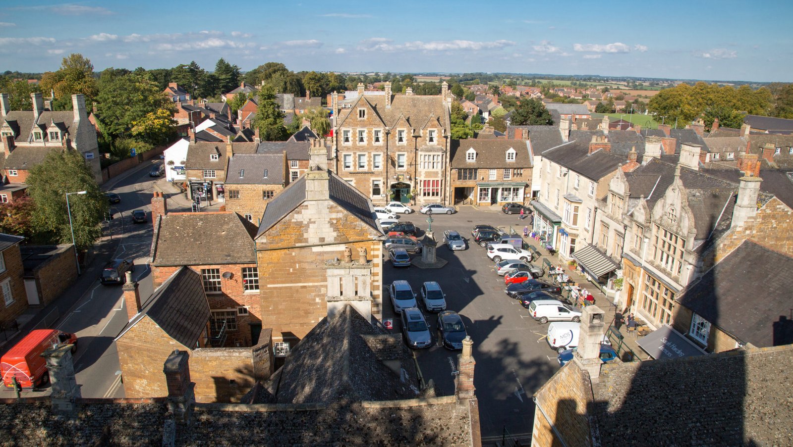 View from the air of a traditional market place. A beautiful coaching in built in stone the colour of gingerbread, pubs, cafes and shops surround parked cars in the centre.