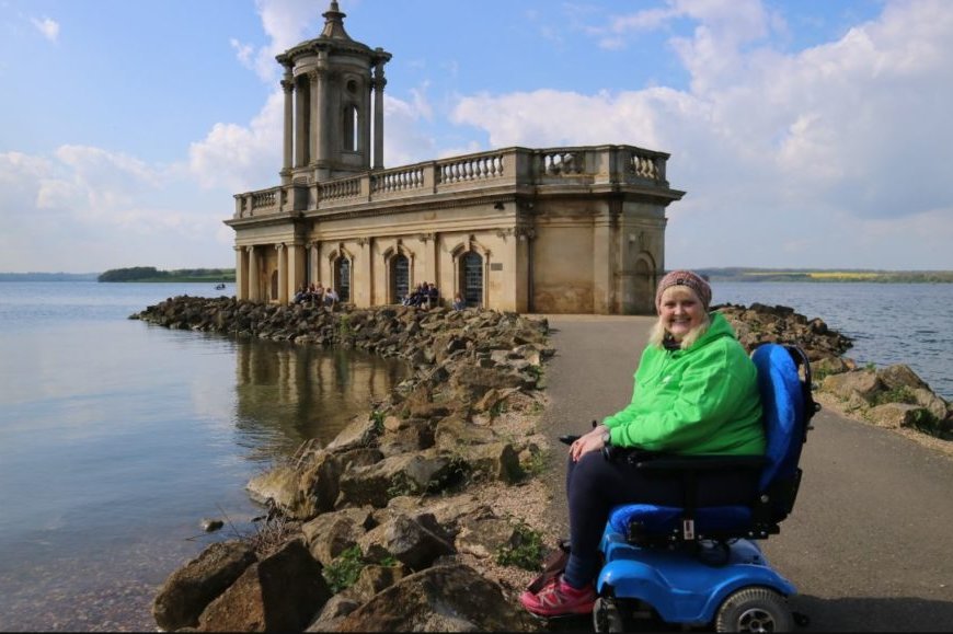 Beautiful blonde haired woman sits in blue powered wheelchair in front of a church with lake behind, blue sky reflects on the water.