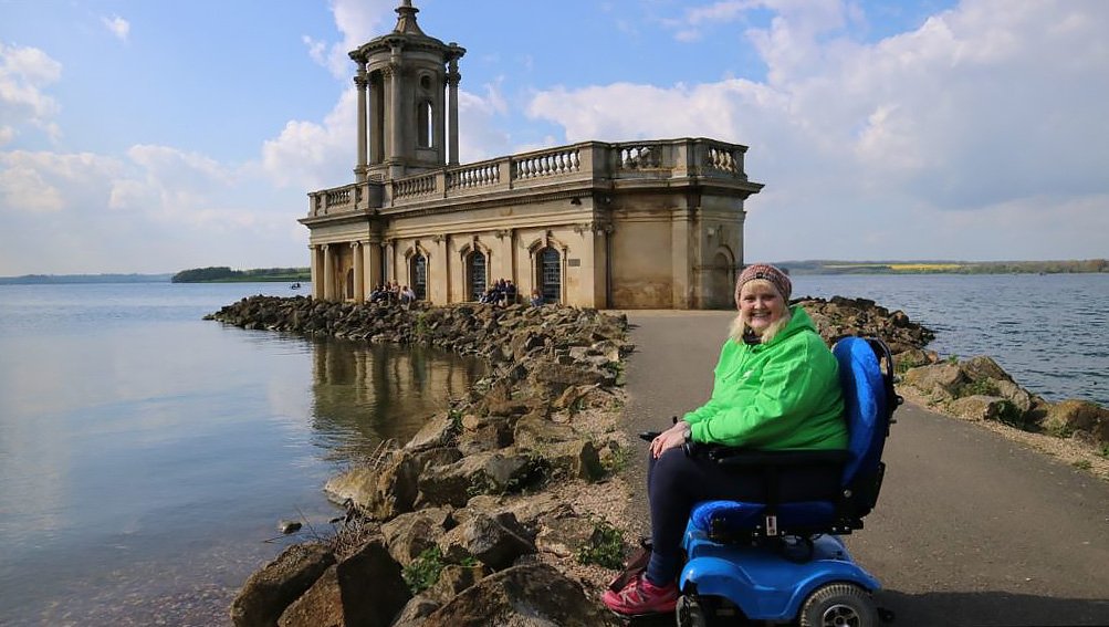 Beautiful blonde haired woman sits in blue powered wheelchair in front of a church with lake behind, blue sky reflects on the water.