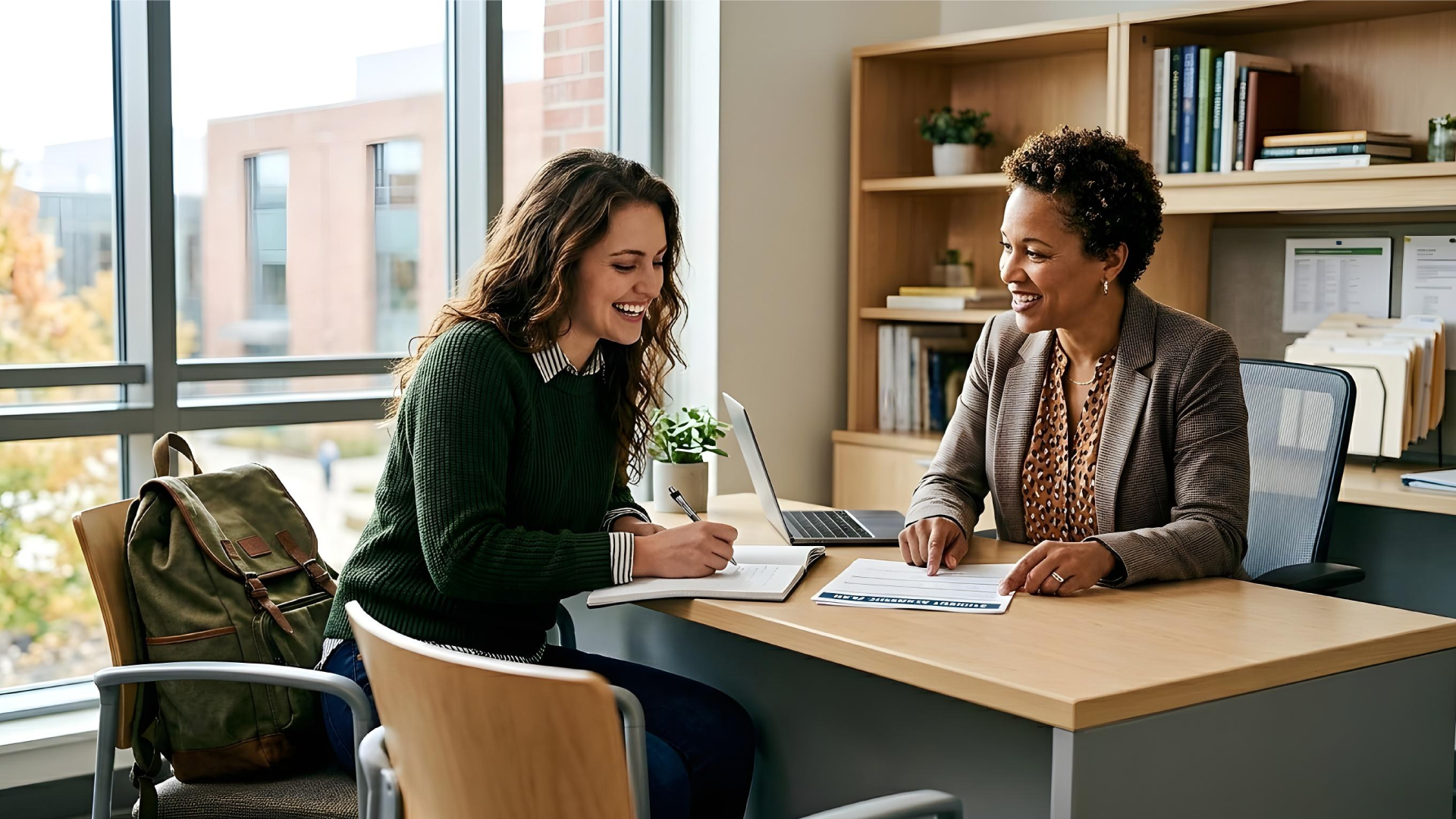 A smiling female university student in a green sweater sits at a desk across from a female advisor in a professional blazer. The student is writing in a notebook while the advisor points to a document. A laptop and bookshelves are on the desk in a bright office with large windows overlooking a campus.