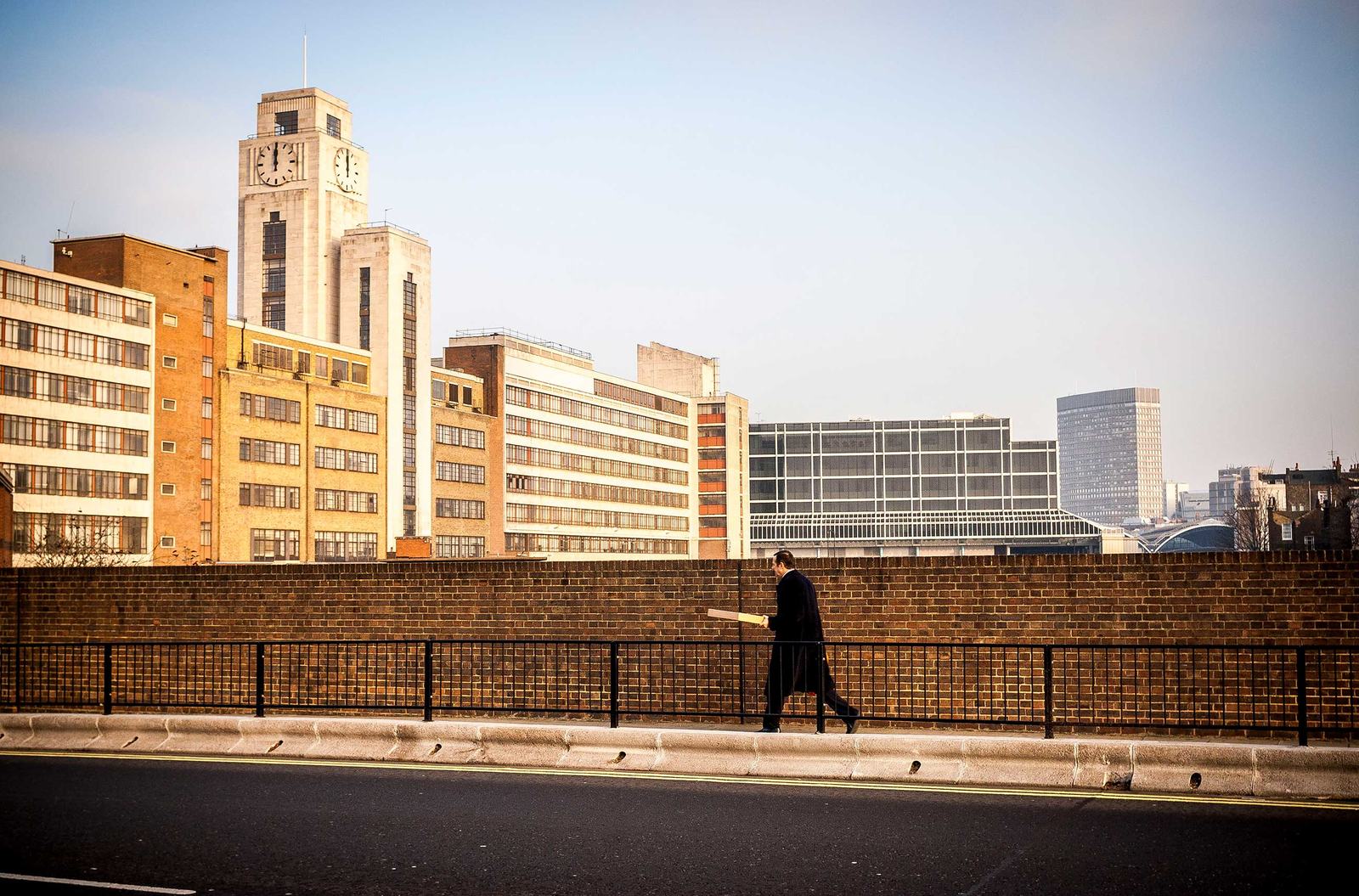 A lone man in a dark coat walks along the pavement beside a brick wall, holding a yellow umbrella; office blocks and a clock tower rise behind under a clear sky.