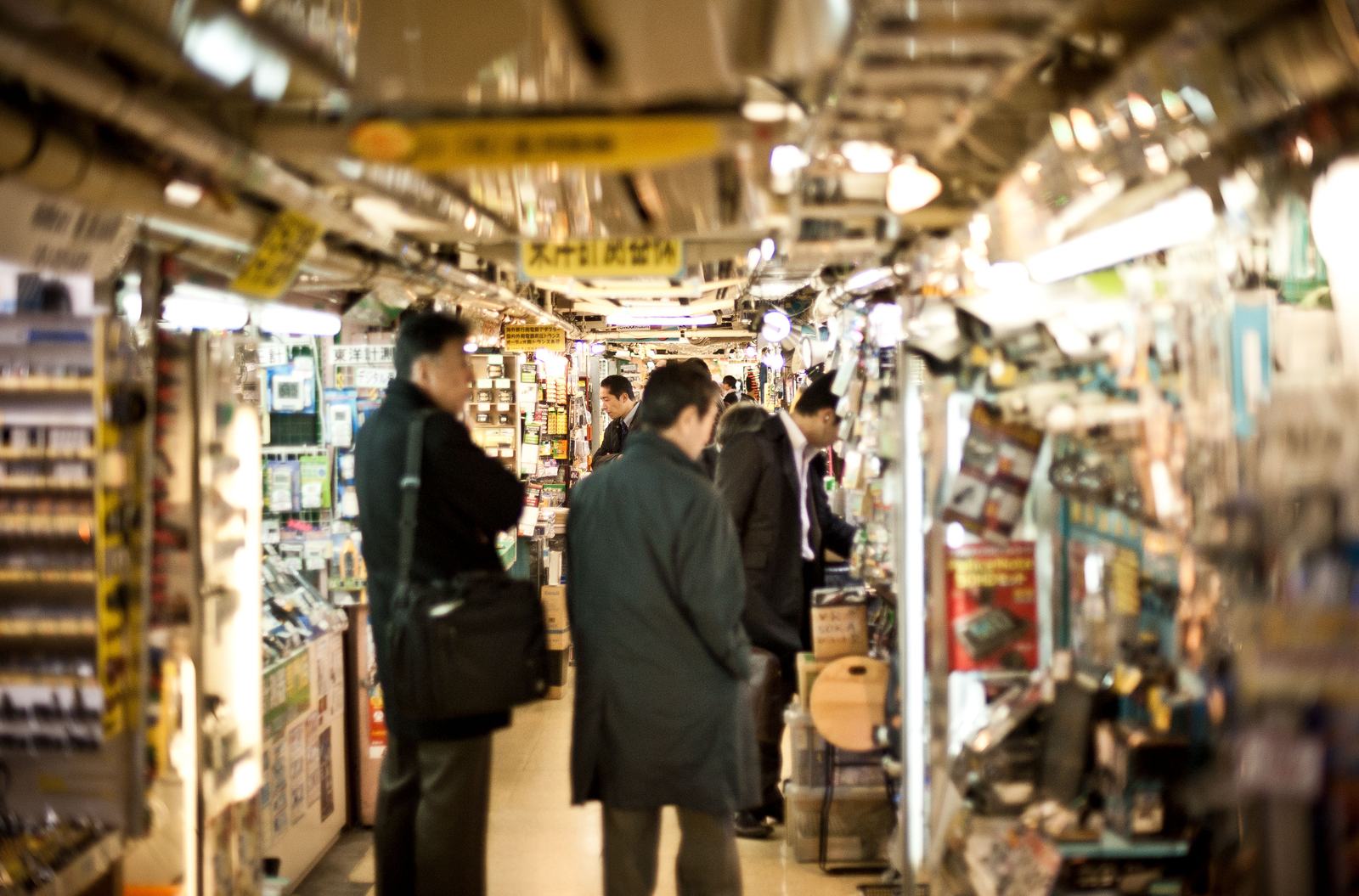 Narrow indoor electronics market aisle with men in coats browsing densely packed stalls of gadgets, components and boxed goods under bright, cluttered lighting.