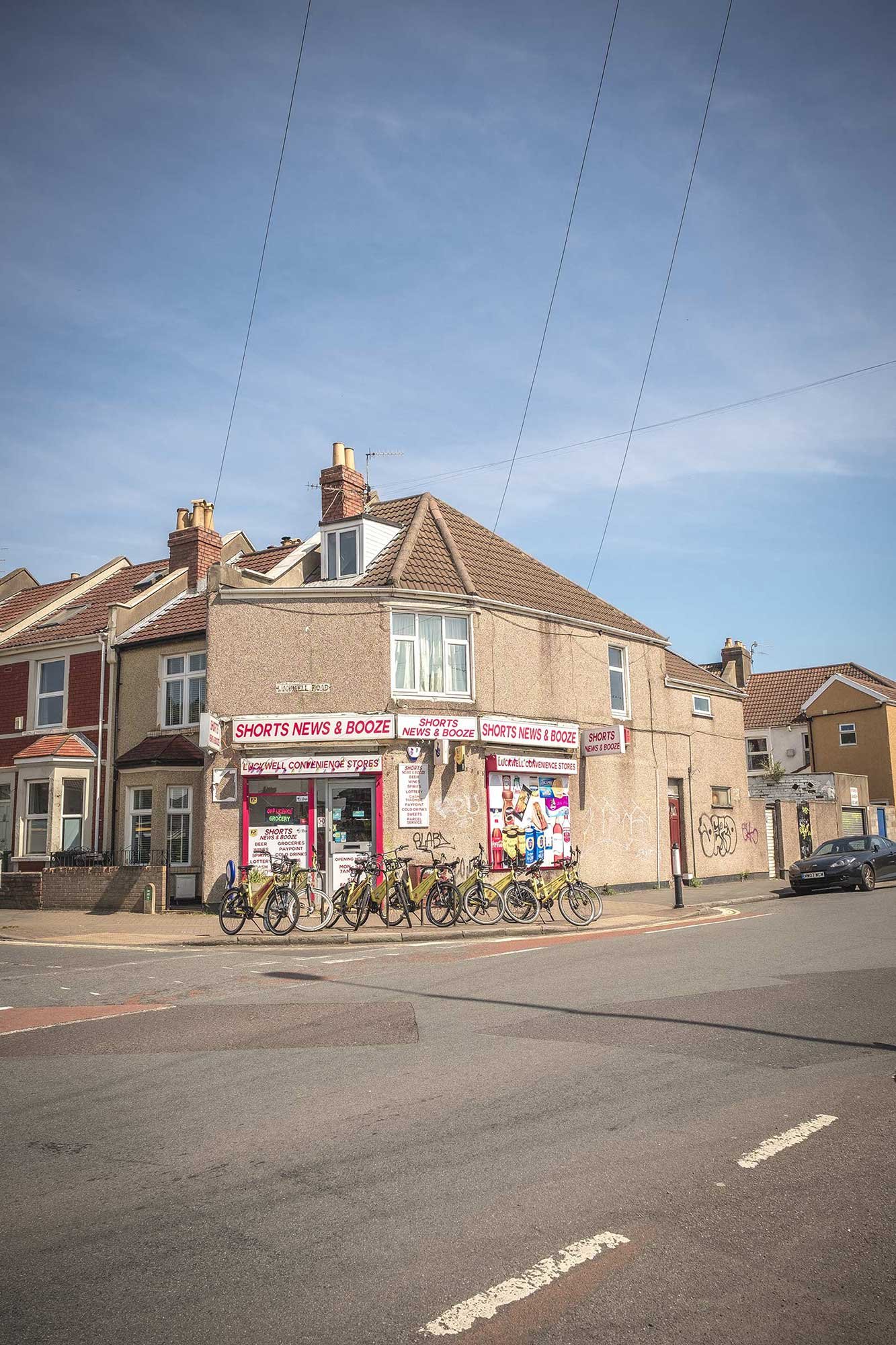 Corner convenience shop 'Shorts News & Booze' in a terraced house, many bikes parked outside, graffiti on the wall, clear blue sky above.