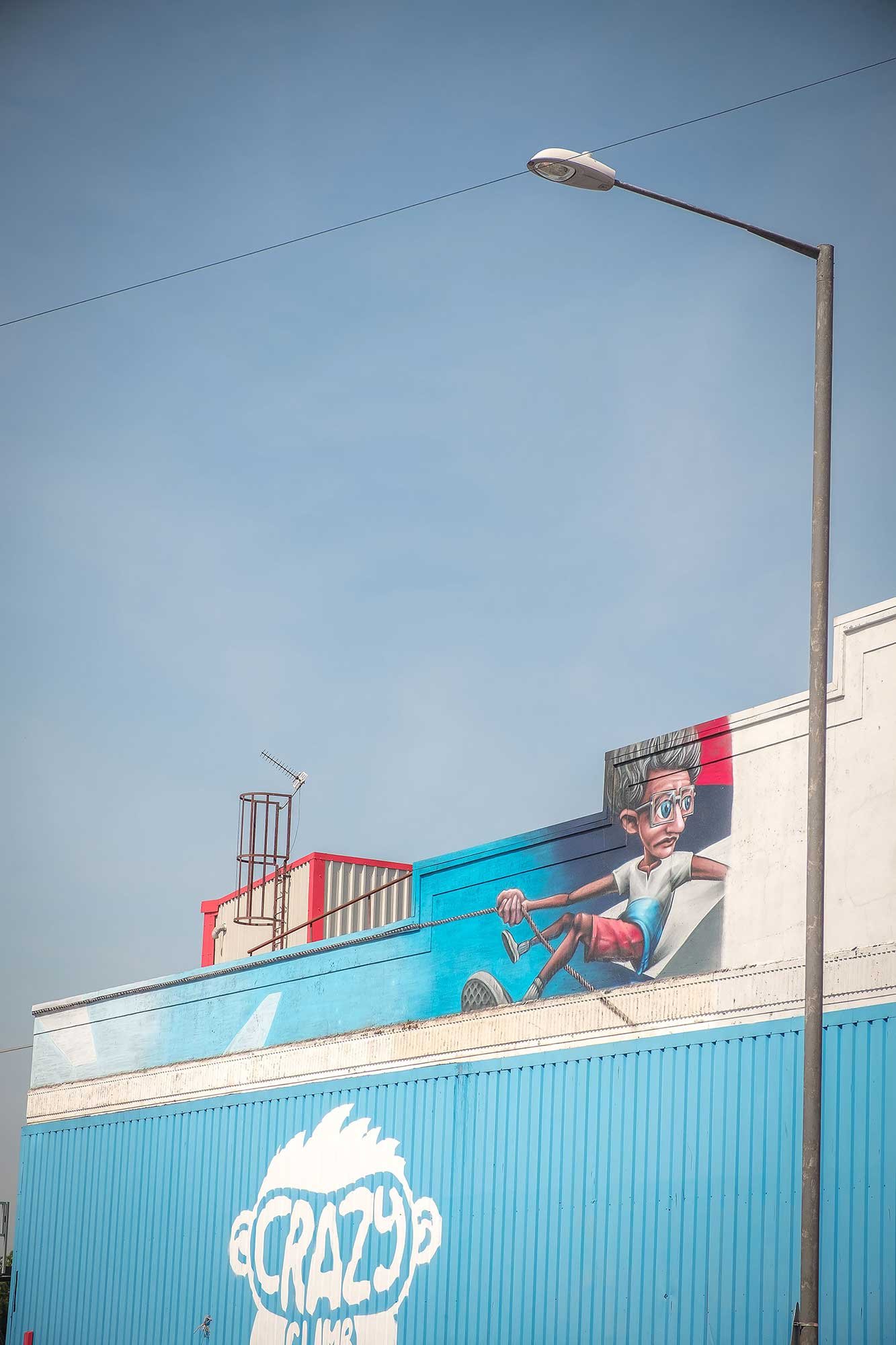 Bright street scene: blue corrugated building with a large mural of a cartoon boy climbing ropes above a 'CRAZY' logo, a tall lamppost and clear sky overhead.