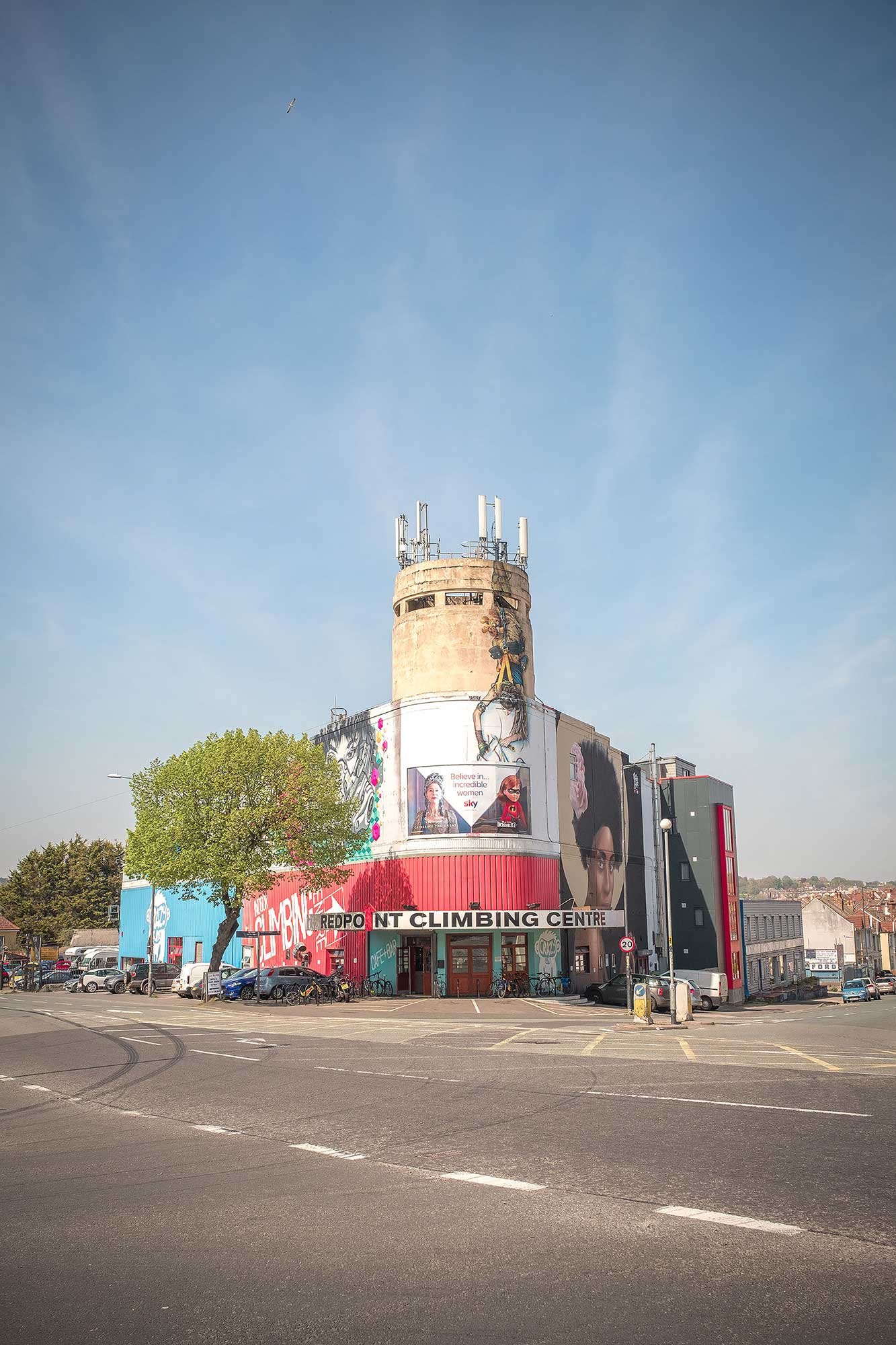 A cylindrical, mural‑covered tower above a red and white building marked "Redpoint Climbing Centre", with a tree, parked cars and a clear blue sky.