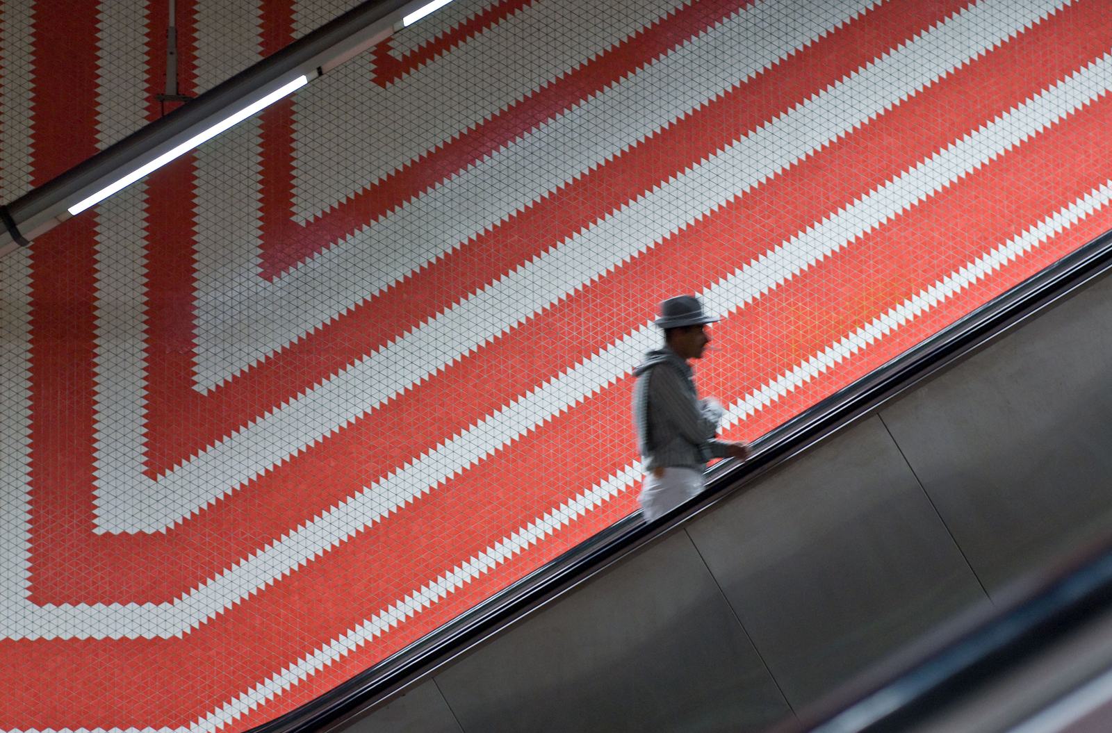 Silhouetted person on an escalator passes a bold red-and-white chevron tiled wall, lit by a fluorescent light.
