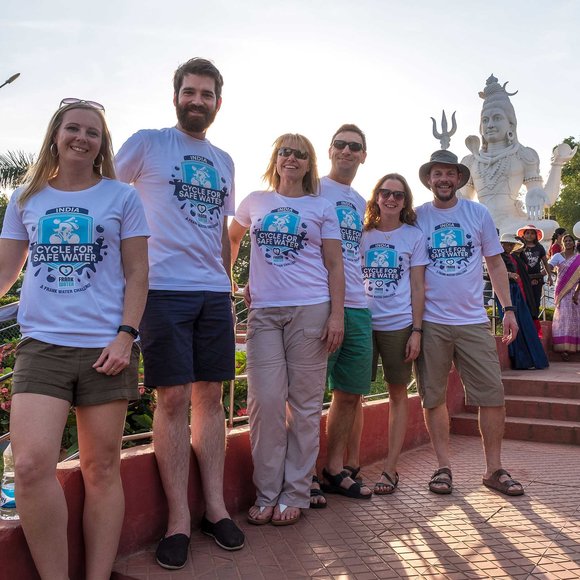 Six adults in matching 'Cycle for Safe Water' T-shirts pose smiling at a sunny temple site, with large white statues of Hindu deities behind them.