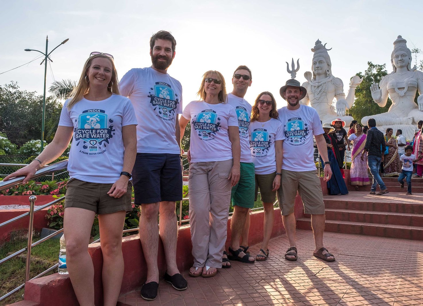 Six adults in matching 'Cycle for Safe Water' T-shirts pose smiling at a sunny temple site, with large white statues of Hindu deities behind them.