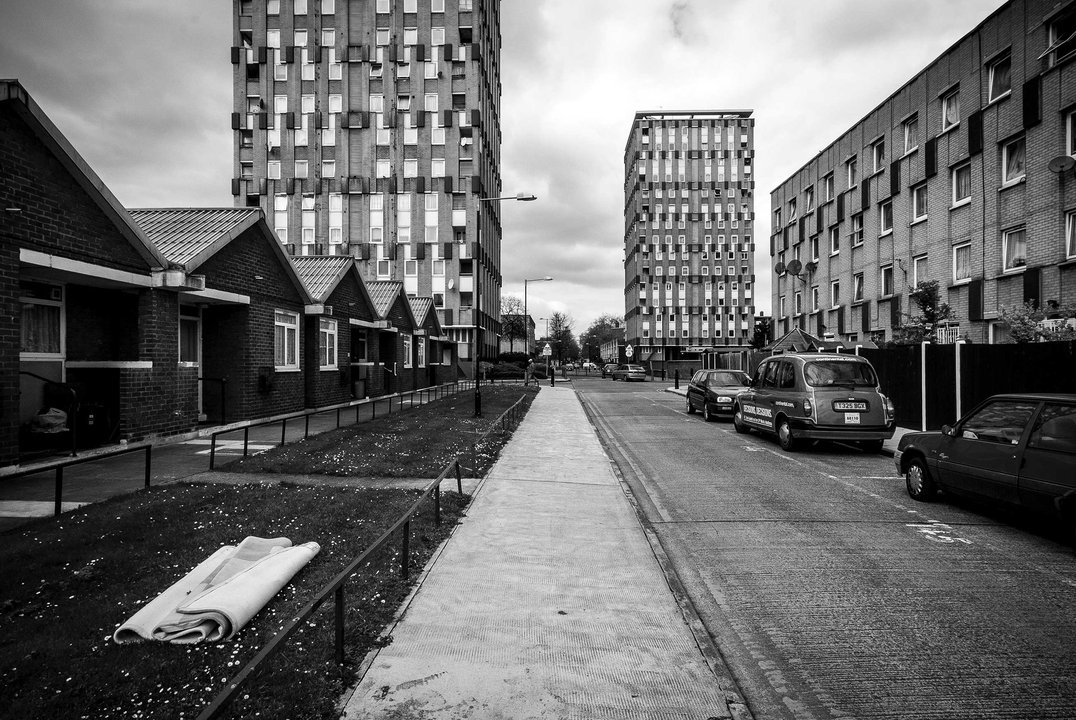 Black-and-white view of a council estate street: low brick bungalows, two high-rise flats ahead, parked cars and a rolled-up carpet on the grass under an overcast sky.