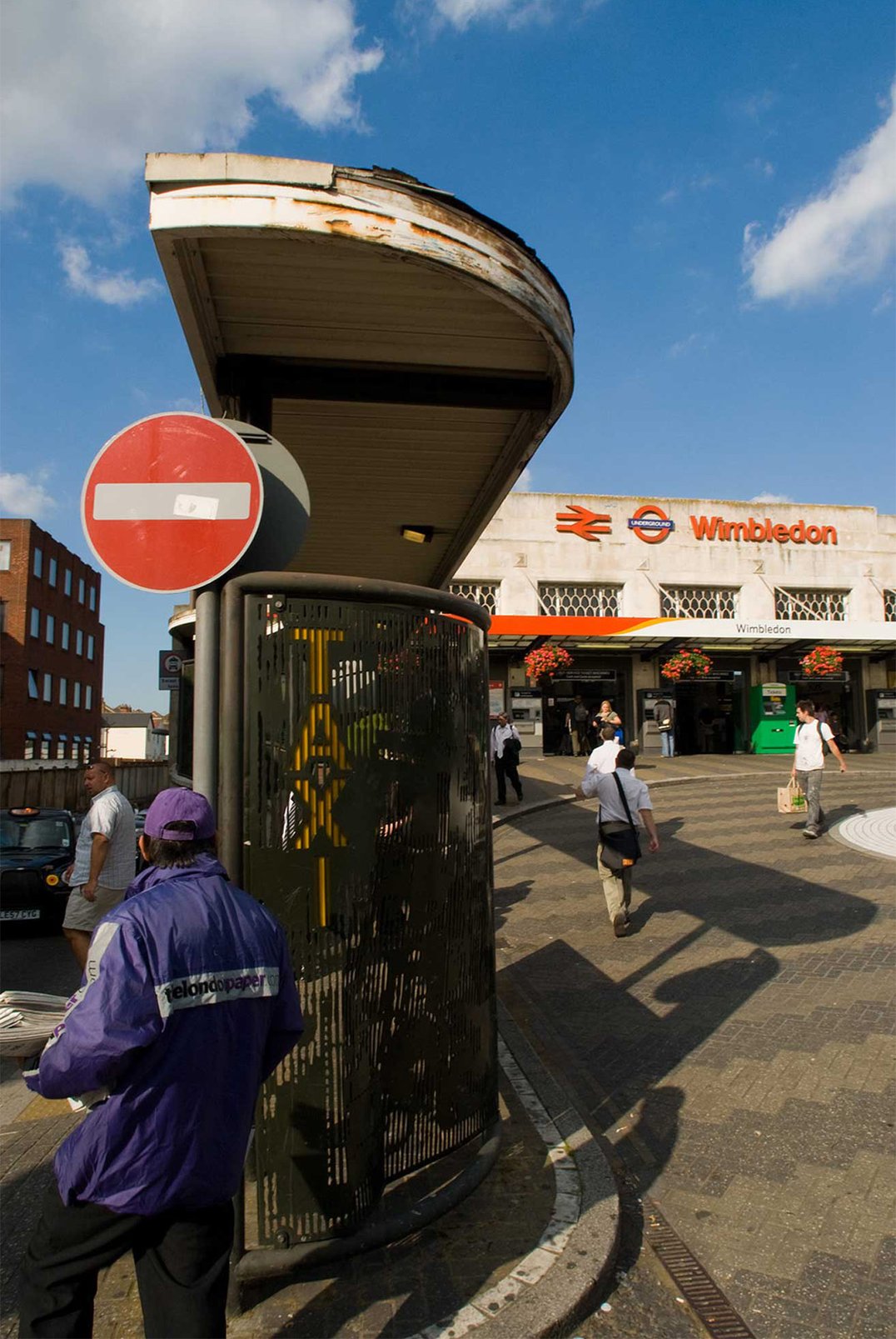Street scene outside Wimbledon station with people, a curved shelter, a no-entry sign, hanging flower baskets and a clear blue sky.