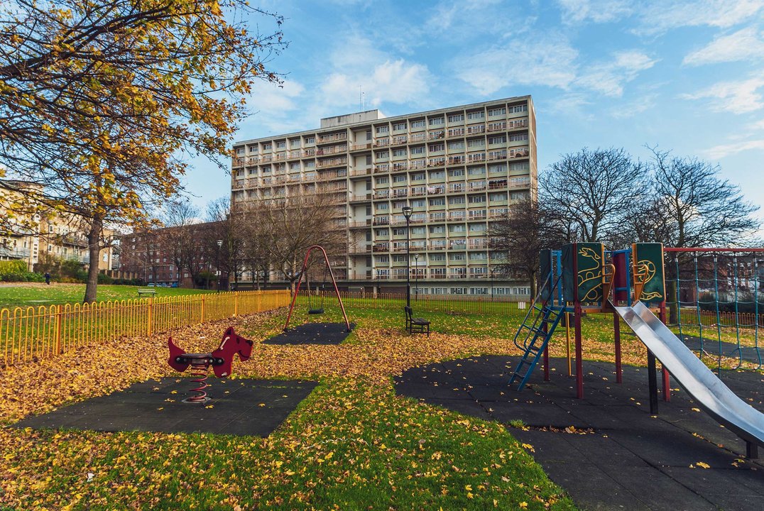 Empty children's playground with a slide, spring rider and swing, carpeted in autumn leaves, set in front of a tall block of flats under a blue sky.