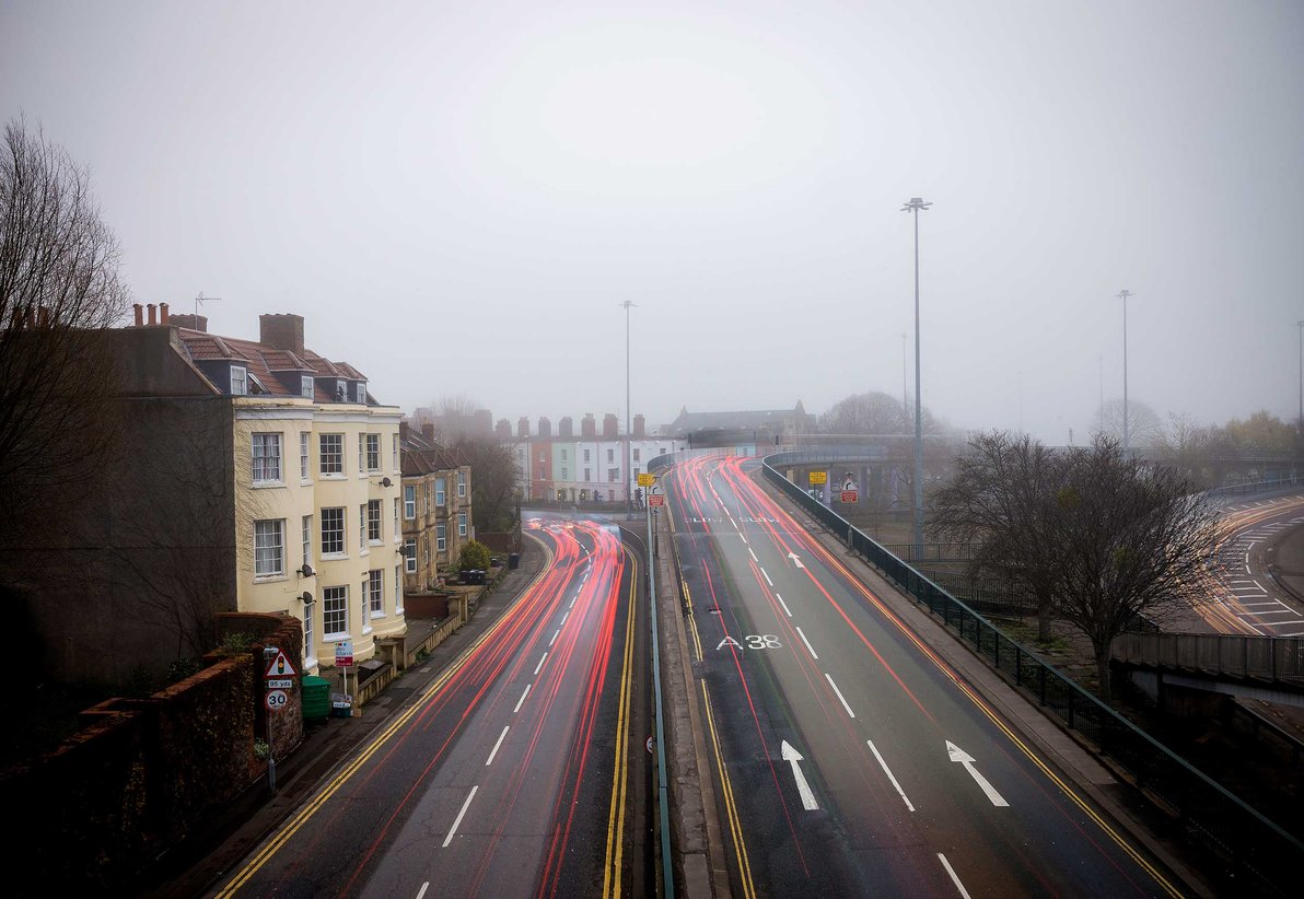 Foggy urban scene: long-exposure light trails on a dual carriageway and flyover, terraced houses left and bare trees right.