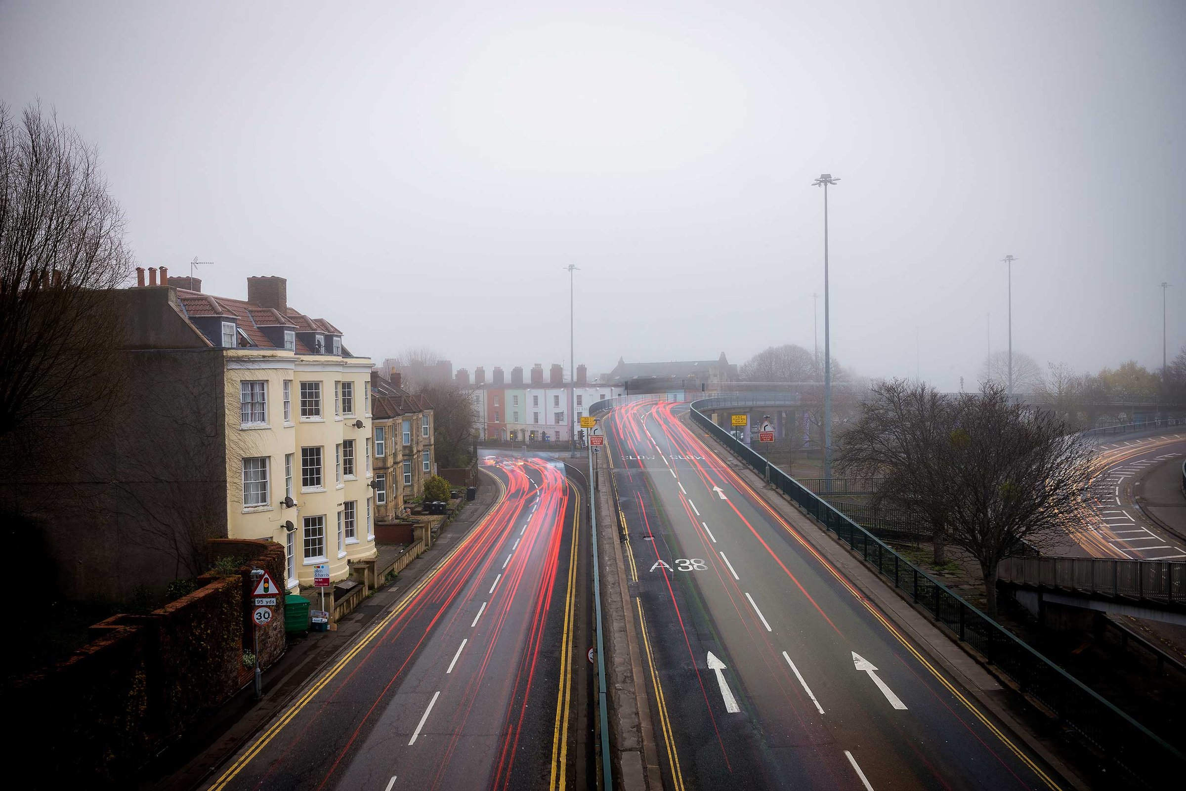 Foggy urban scene: long-exposure light trails on a dual carriageway and flyover, terraced houses left and bare trees right.