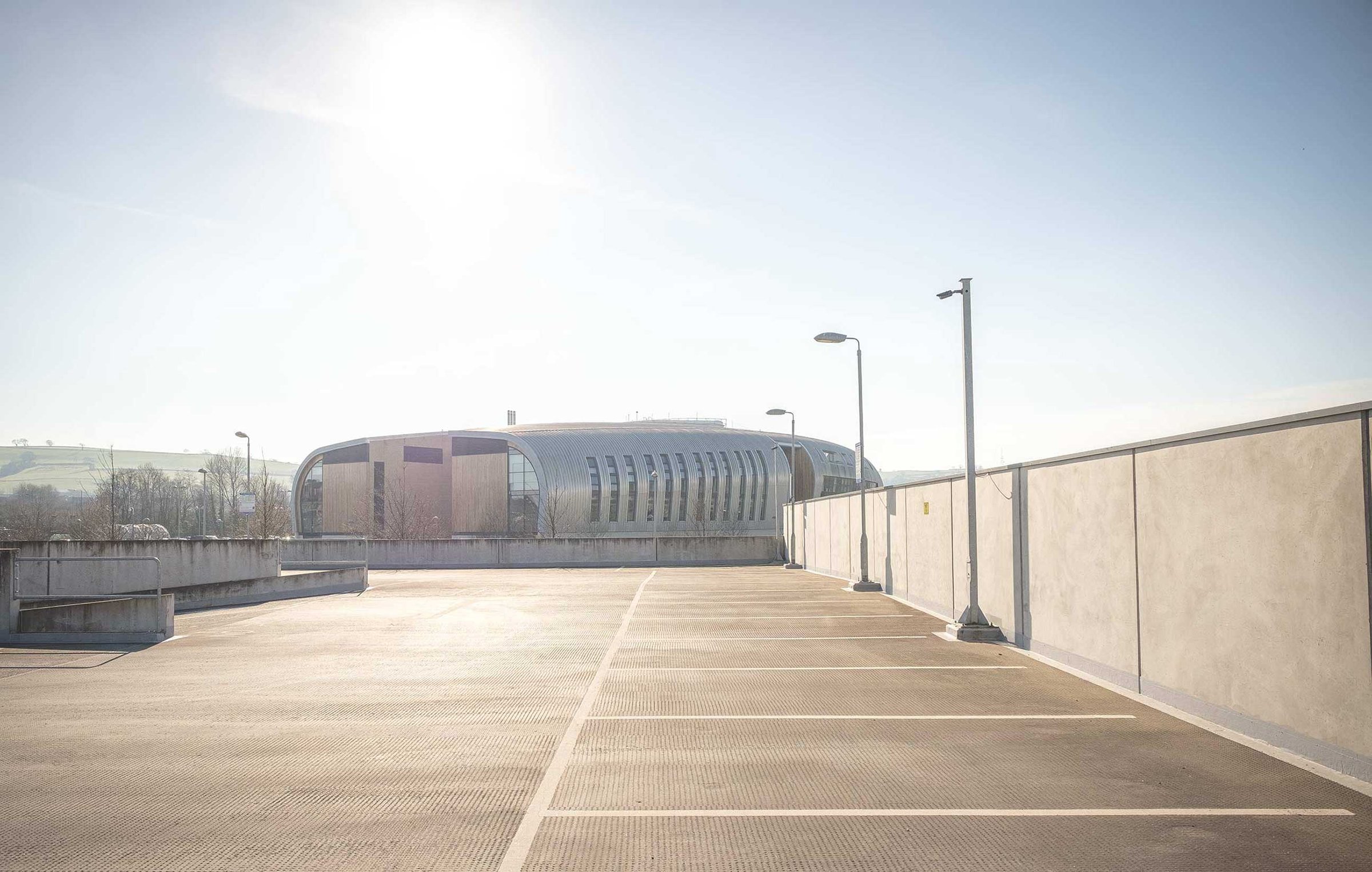 Empty rooftop car park under bright winter sun, lined bays and lampposts, concrete walls; a modern curved building and rolling hills visible in the hazy distance.