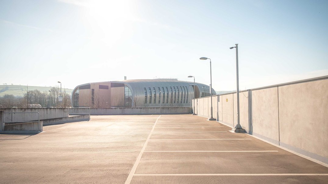 Empty rooftop car park bathed in soft morning light, lined with lamp posts; a modern curved building and distant rolling hills beyond a low concrete wall.