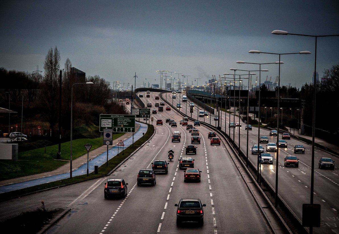Wet multi-lane urban dual carridgeway with traffic heading over a bridge towards London, lit streetlights, a blue cycle lane and an overcast sky.