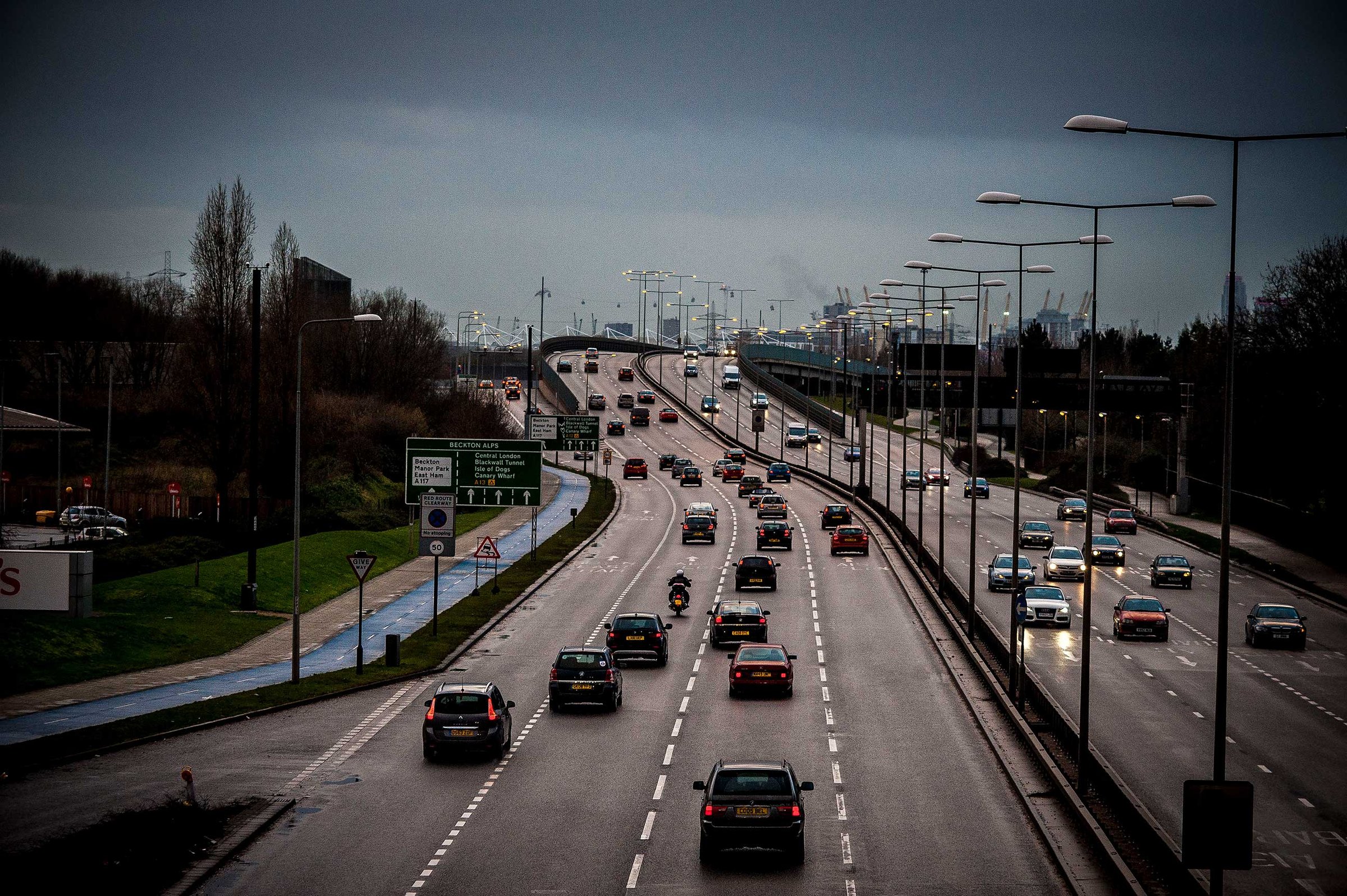 Wet multi-lane urban dual carridgeway with traffic heading over a bridge towards London, lit streetlights, a blue cycle lane and an overcast sky.