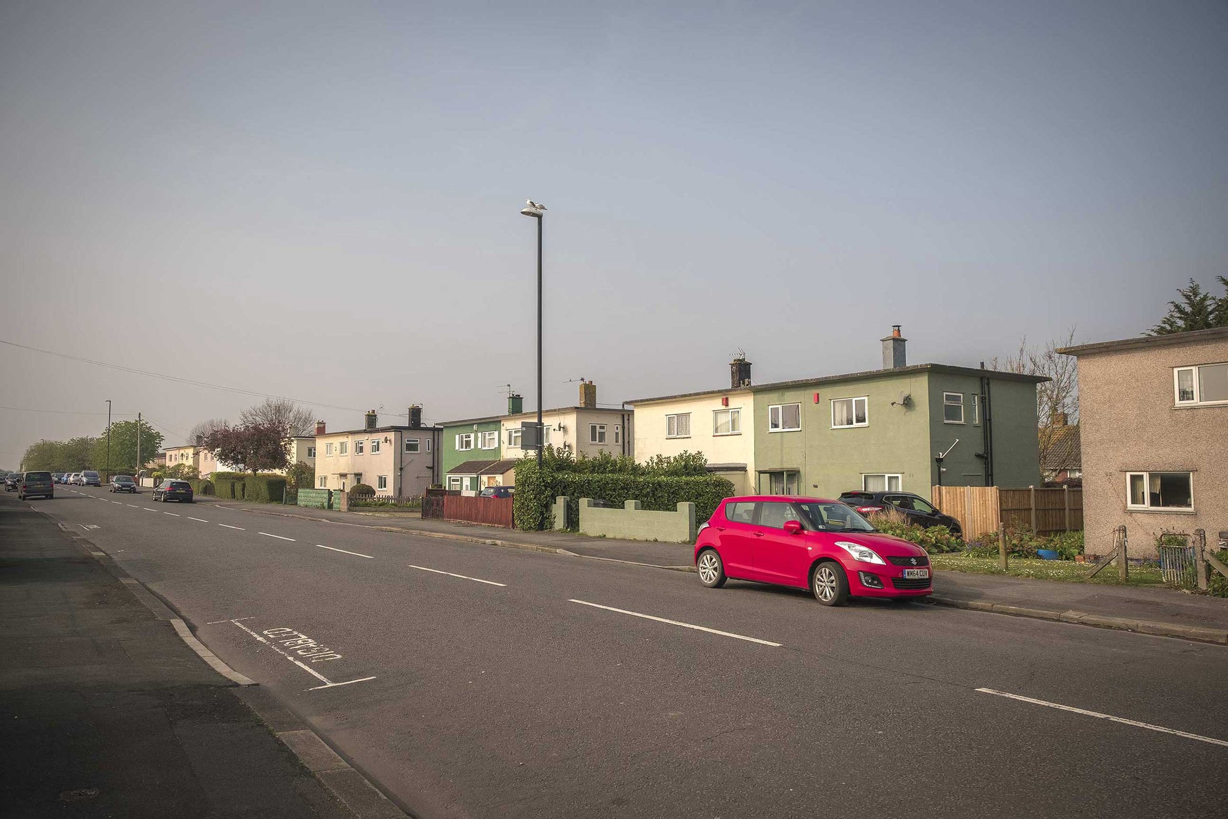 Quiet suburban street with rows of low‑rise semi‑detached houses, a tall lamppost and a bright red hatchback parked at the kerb under a pale, overcast sky.