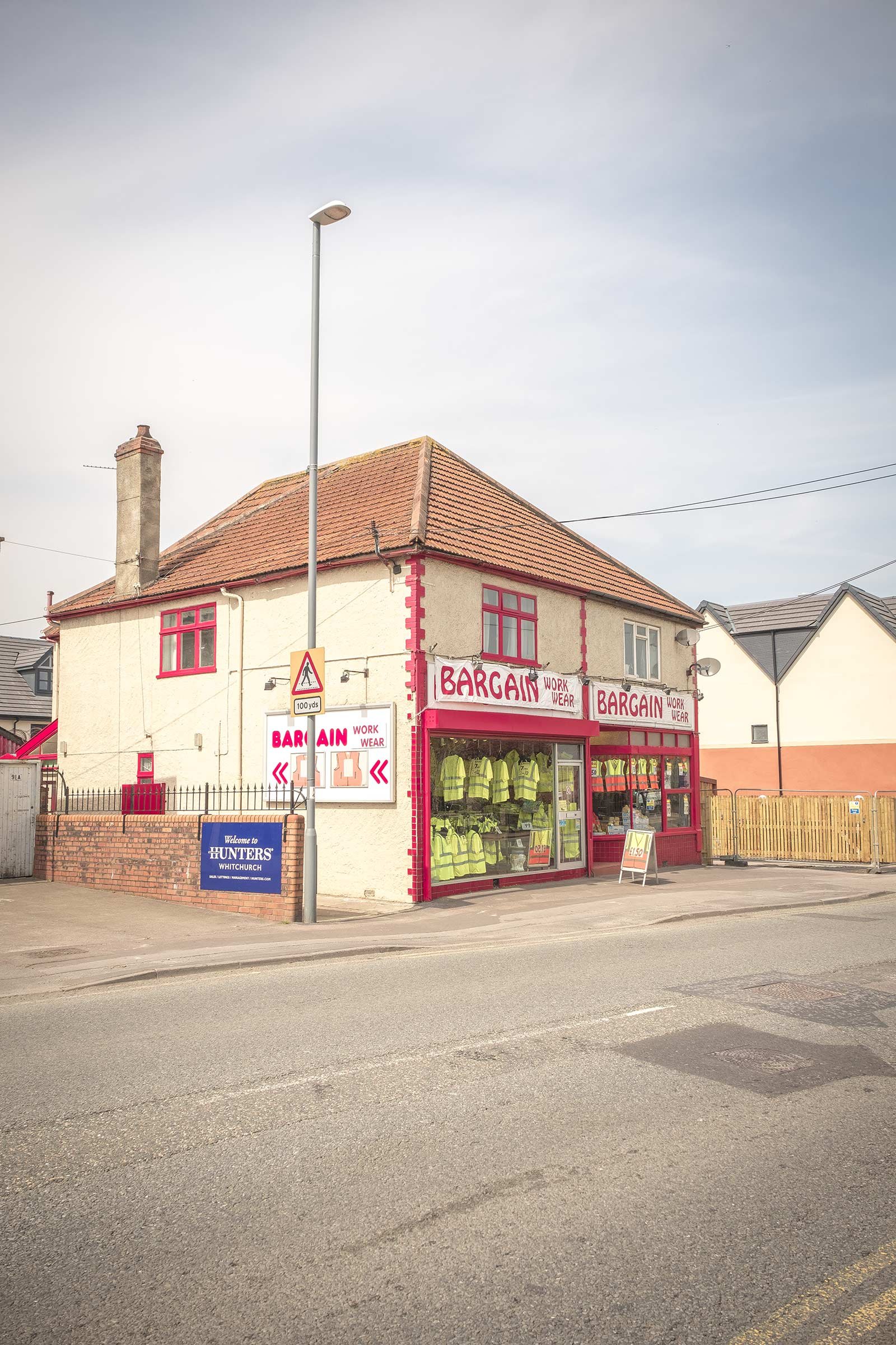Corner shop 'Bargain Workwear' with red trim and a window display of high-visibility jackets, cream walls and tiled roof on a quiet street beneath a pale sky.