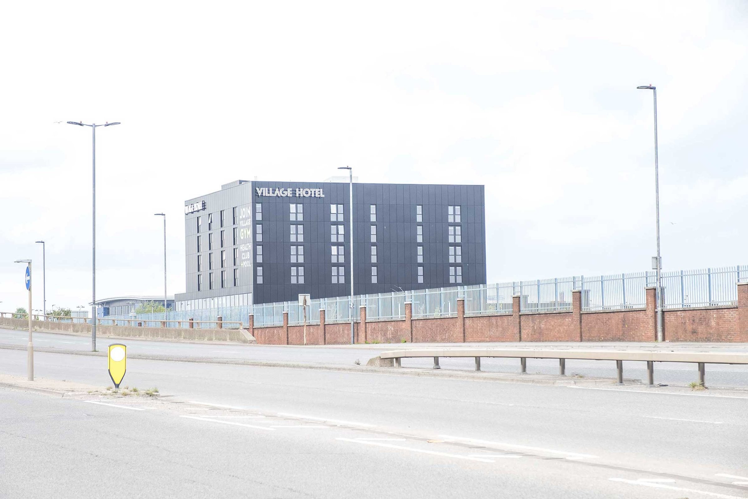 Modern dark 'Village Hotel' block behind a brick wall beside a quiet multi‑lane road with lampposts and a central refuge.