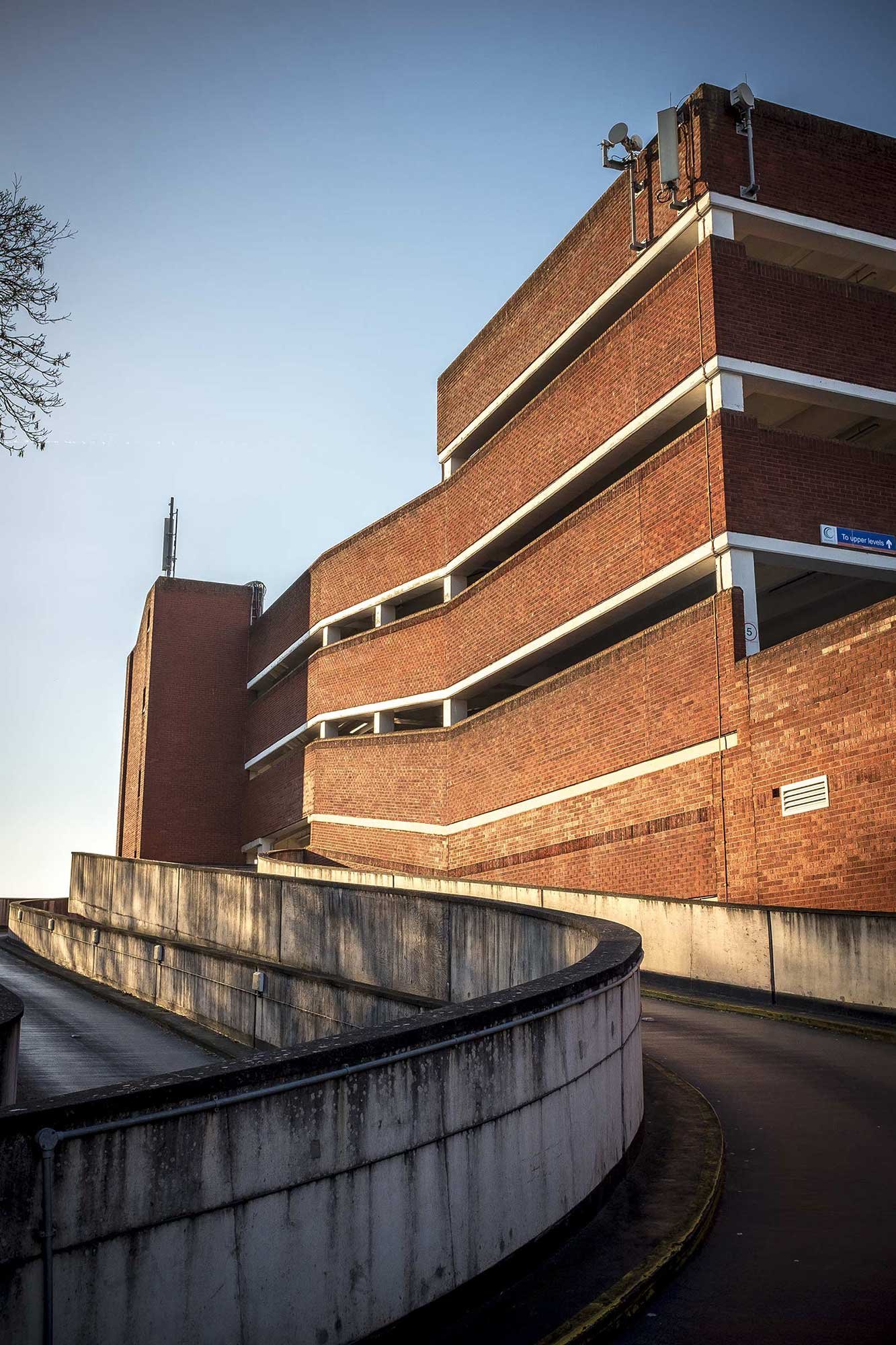 Curving concrete ramp leading to a multi-storey red-brick car park with white bands, bathed in low sunlight under a clear sky.