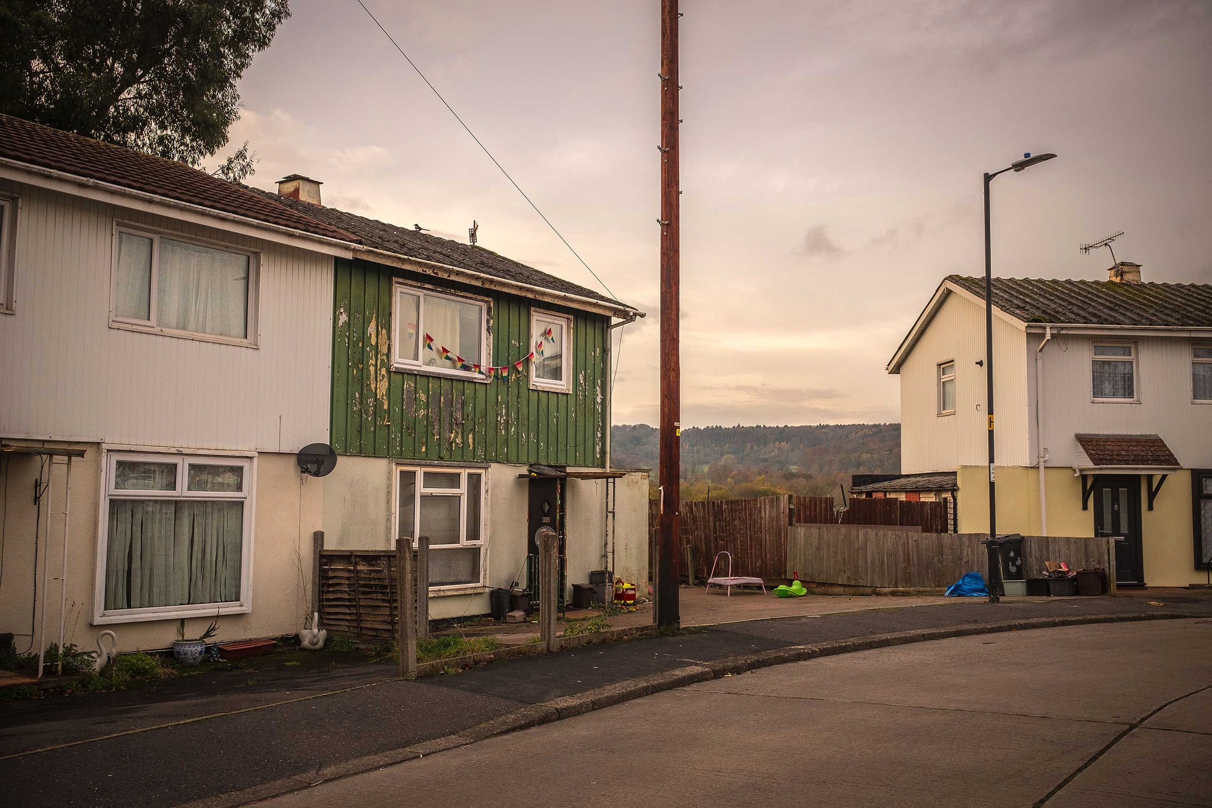 Quiet suburban street at dusk with semi-detached houses, one with weathered green cladding and bunting. Lamppost, bins and children's toys sit by a fenced yard, hills beyond.