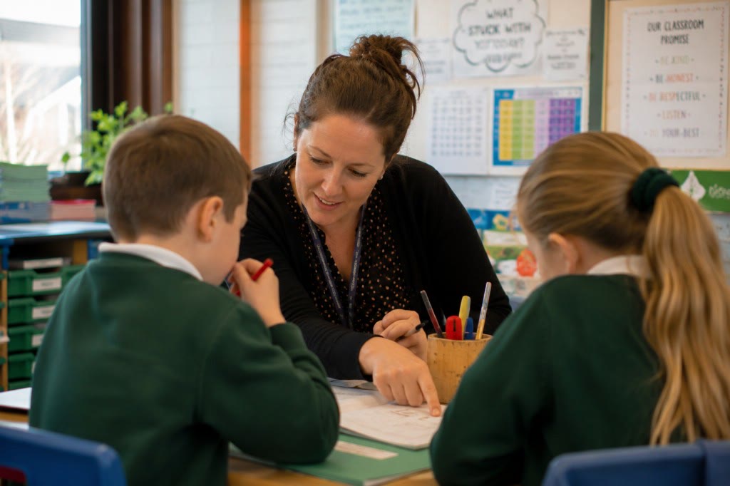 Teacher helping two pupils with their work at a desk, photographed from behind the pupils