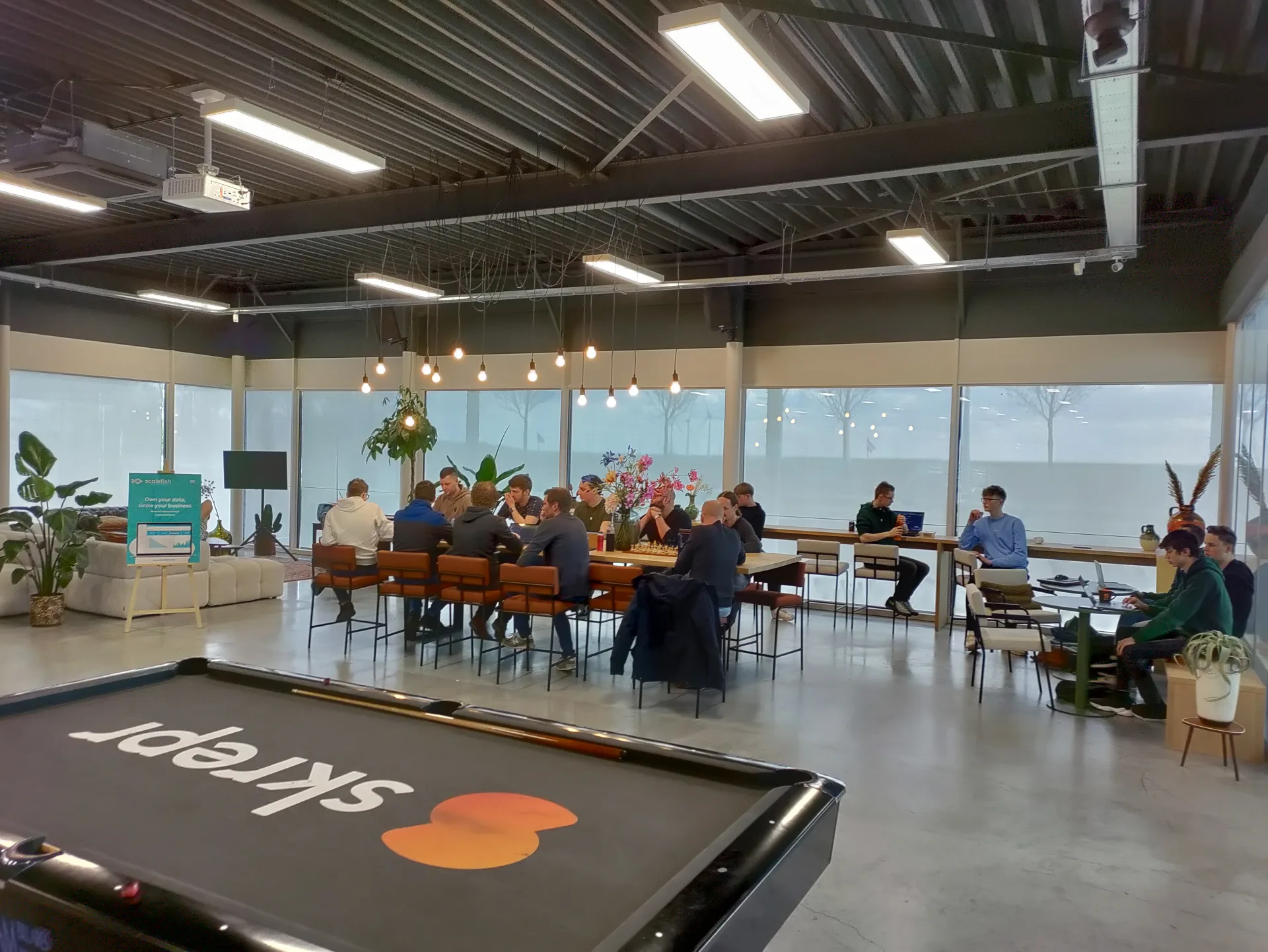 Skrepr-team verzameld in de gezamenlijke ruimte, collega's sparren aan lange tafels, biljart op de voorgrond met Skrepr-logo