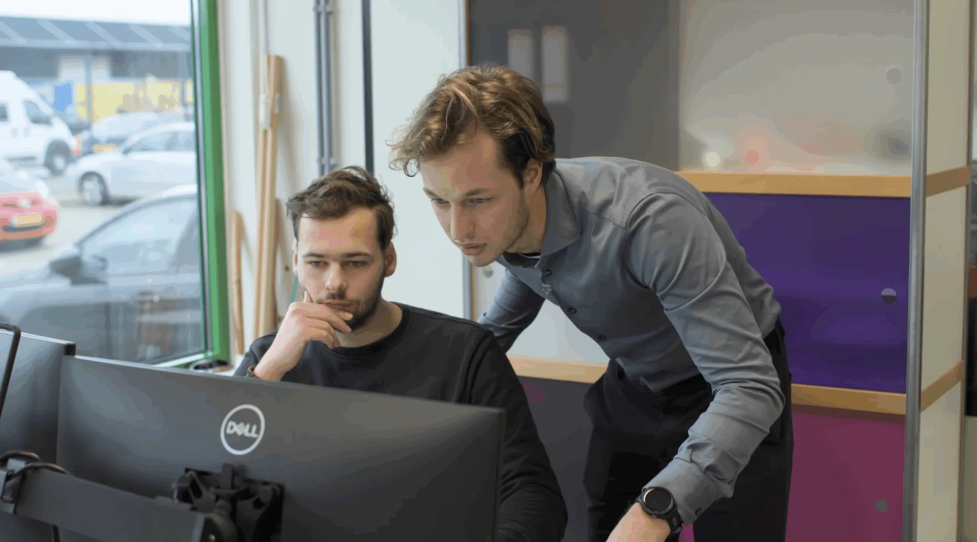 Twee collega's buigen zich over een Dell-monitor en bespreken live productie-data of een AI-agent in uitvoering