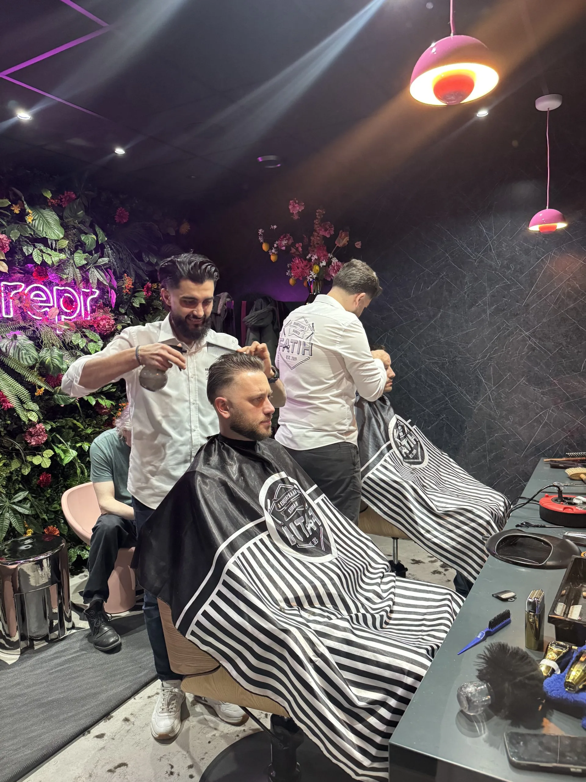 Twee kappers werken zorgvuldig aan klanten in een stijlvolle barbershop met neonverlichting en plantenwand