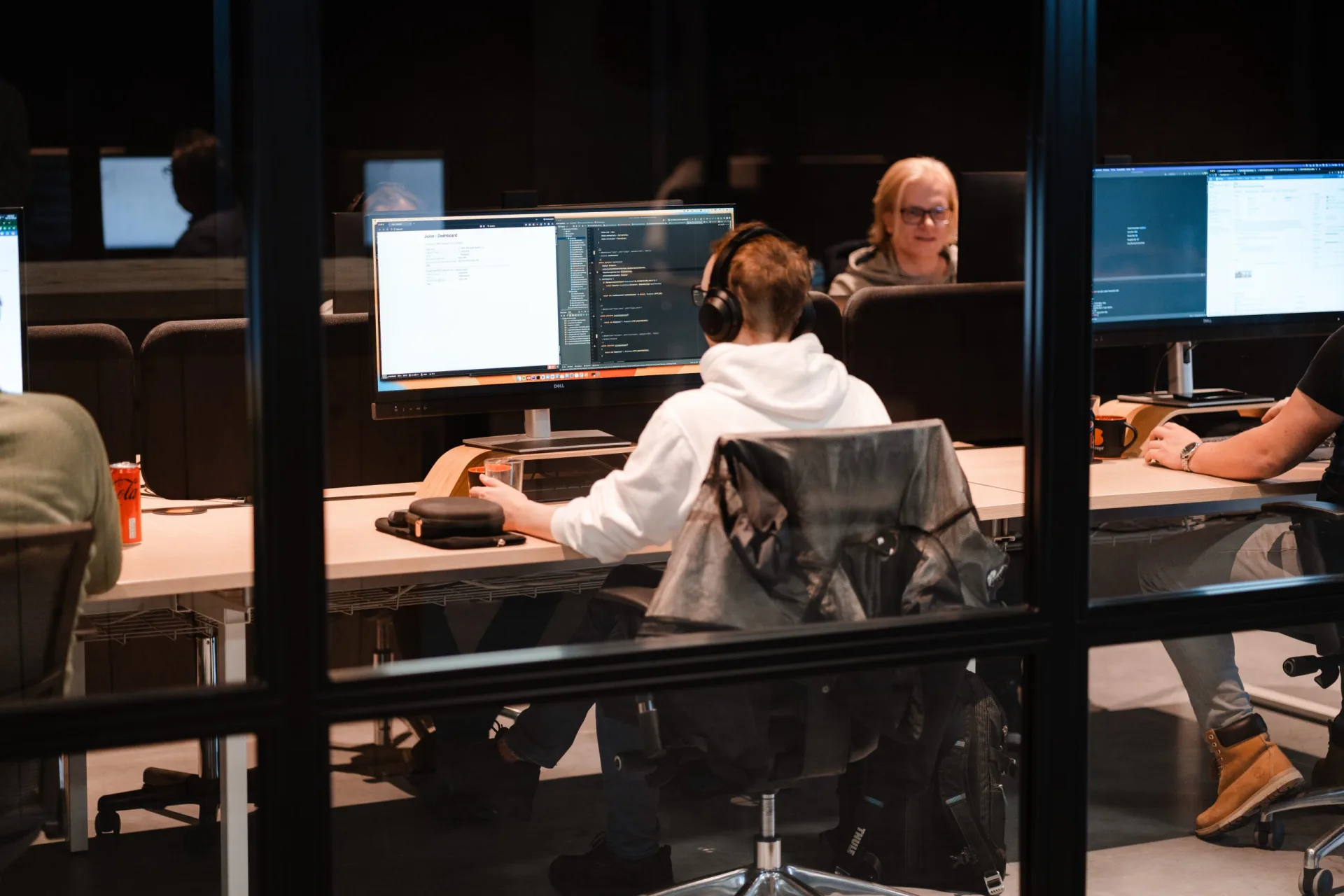 Developer met koptelefoon werkt aan code-editor op groot scherm, collega's actief op de achtergrond in open kantoorruimte