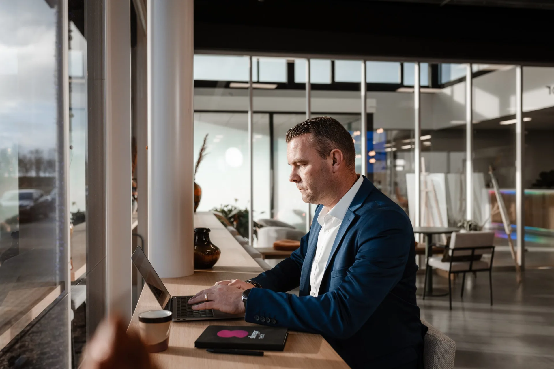 Collega werkt geconcentreerd aan laptop in lichte kantoorruimte met uitzicht, koffie binnen handbereik