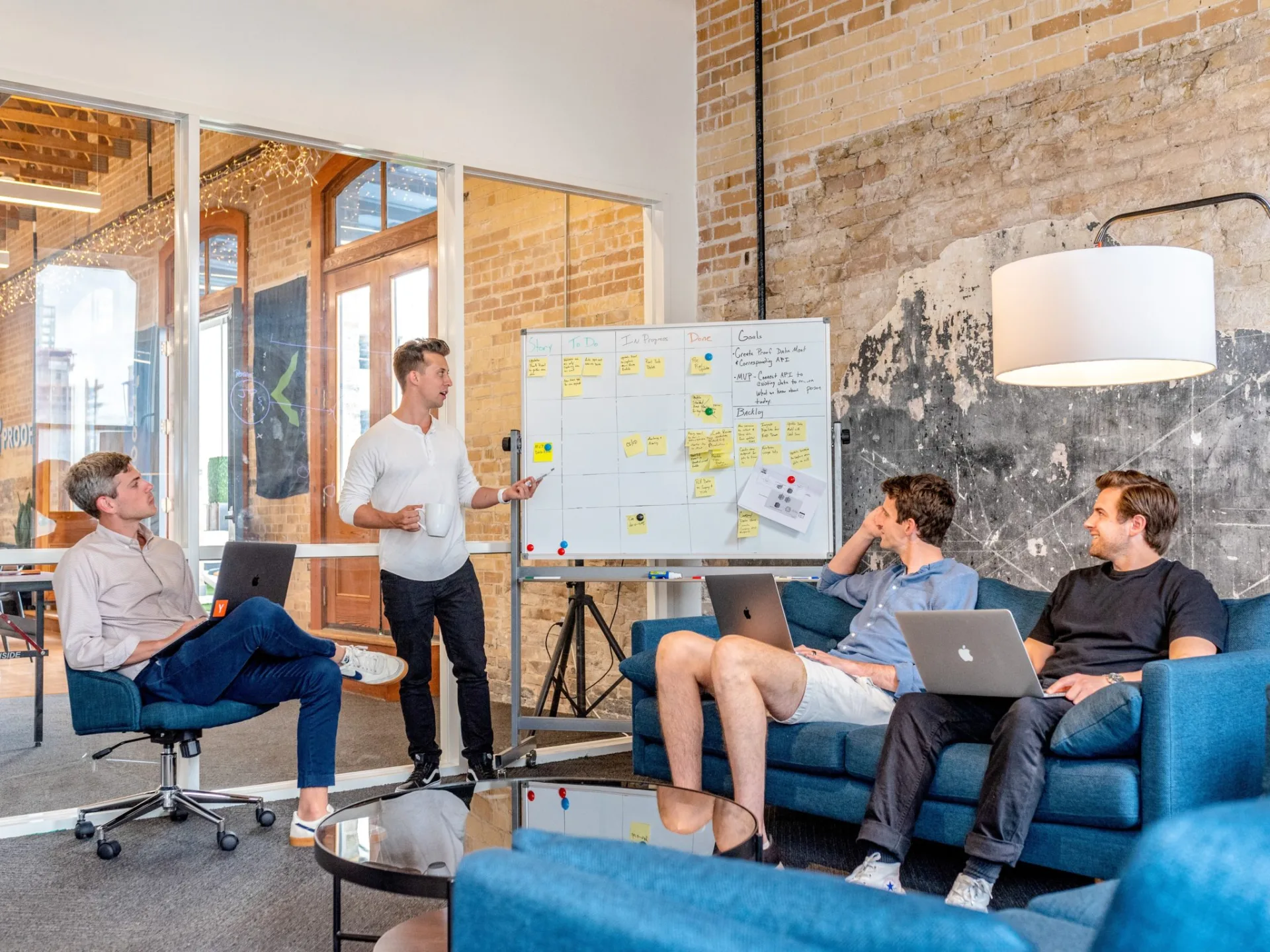 Vier collega's in een informele sessie rond een whiteboard met sticky notes, twee met laptop, één presenteert staand