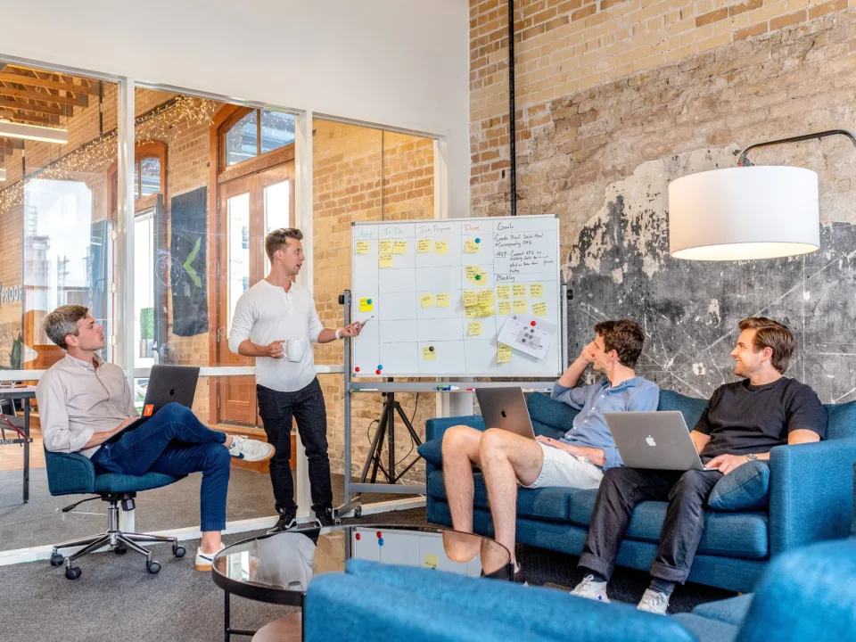 Vier collega's in een informele sessie rond een whiteboard met sticky notes, twee met laptop, één presenteert staand
