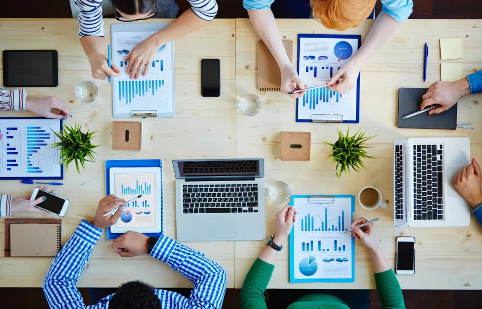 Team van zes collega's analyseert productie-dashboards op laptops, tablets en clipboards rond een vergadertafel