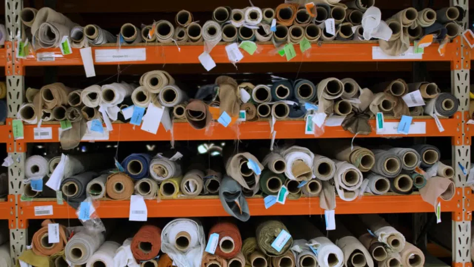 Magazijn met honderden opgerolde stoffen op oranje stellingkasten, voorzien van kleurgecodeerde labels en barcodes voor voorraadregistratie