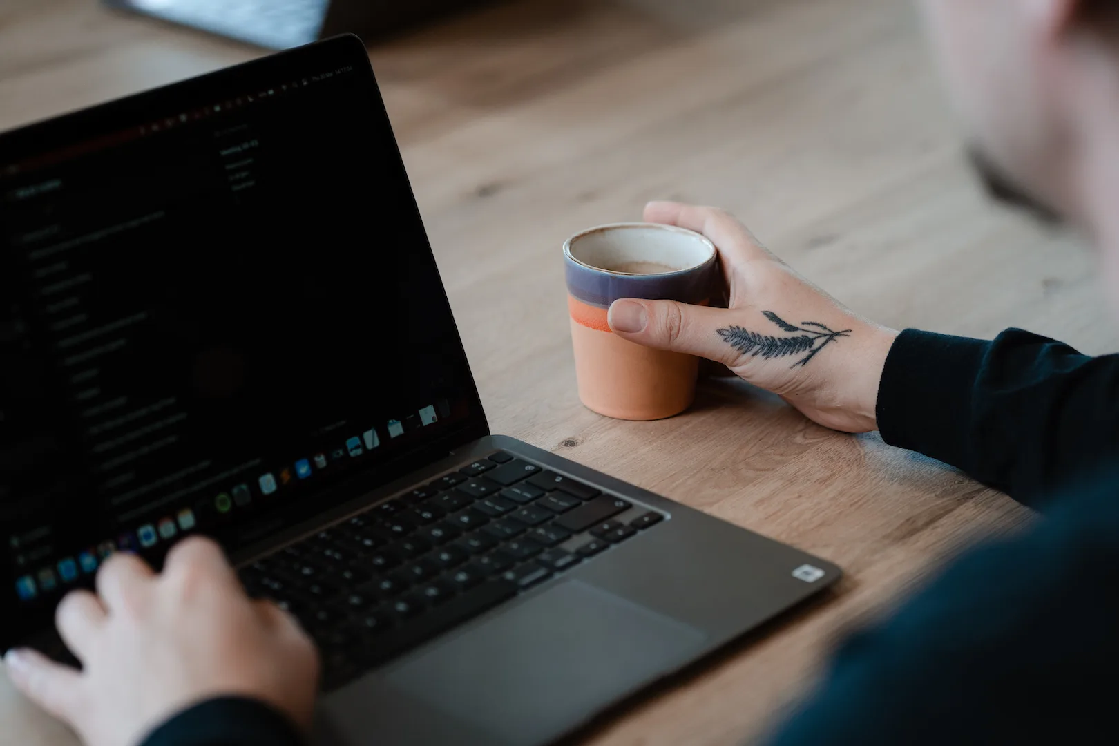 Developer houdt koffiekopje vast en werkt aan dashboard op MacBook, tatoeage op hand zichtbaar