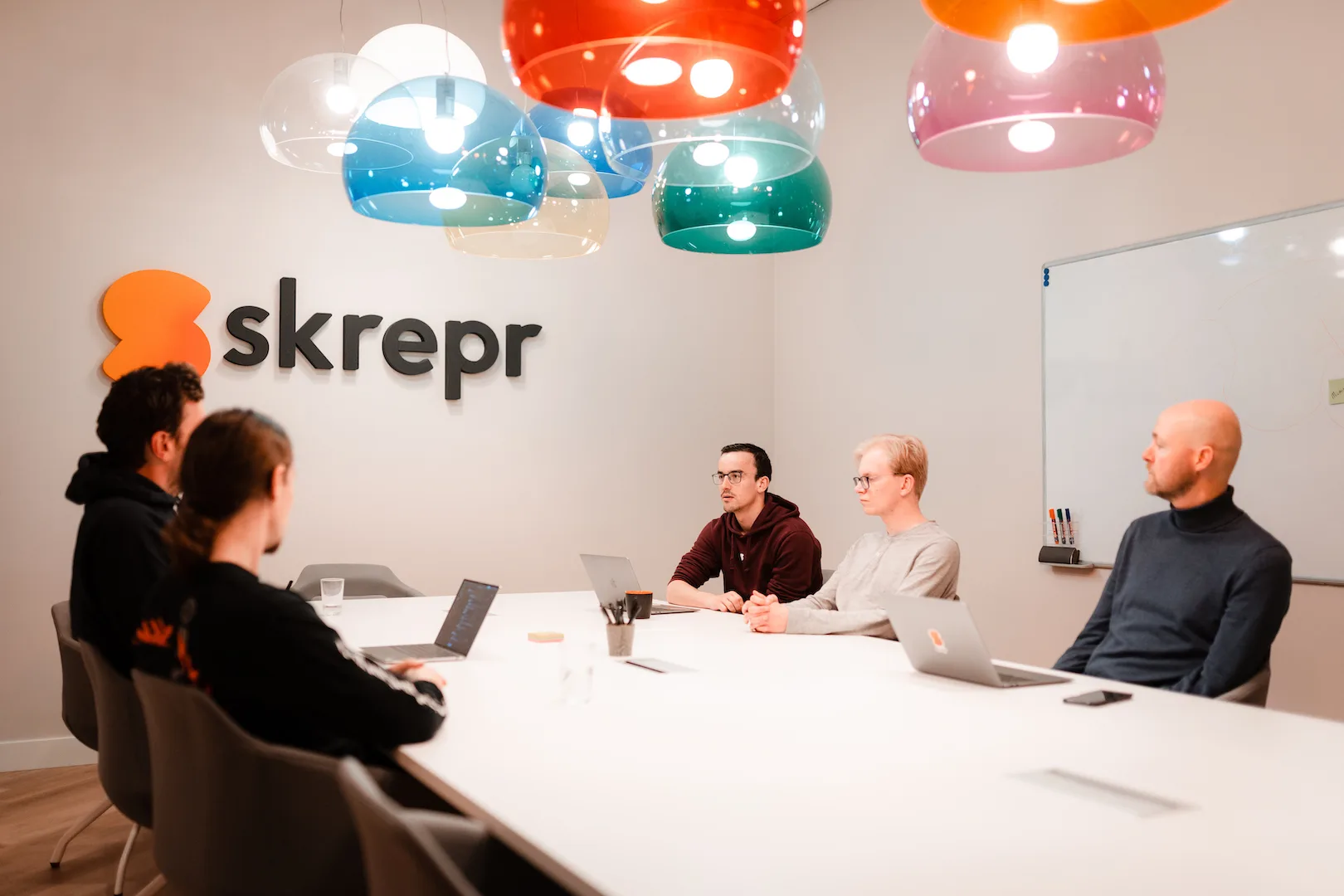 Skrepr-team in vergaderzaal rond een tafel met laptops, bezig met een productsessie onder kleurrijke hanglampen